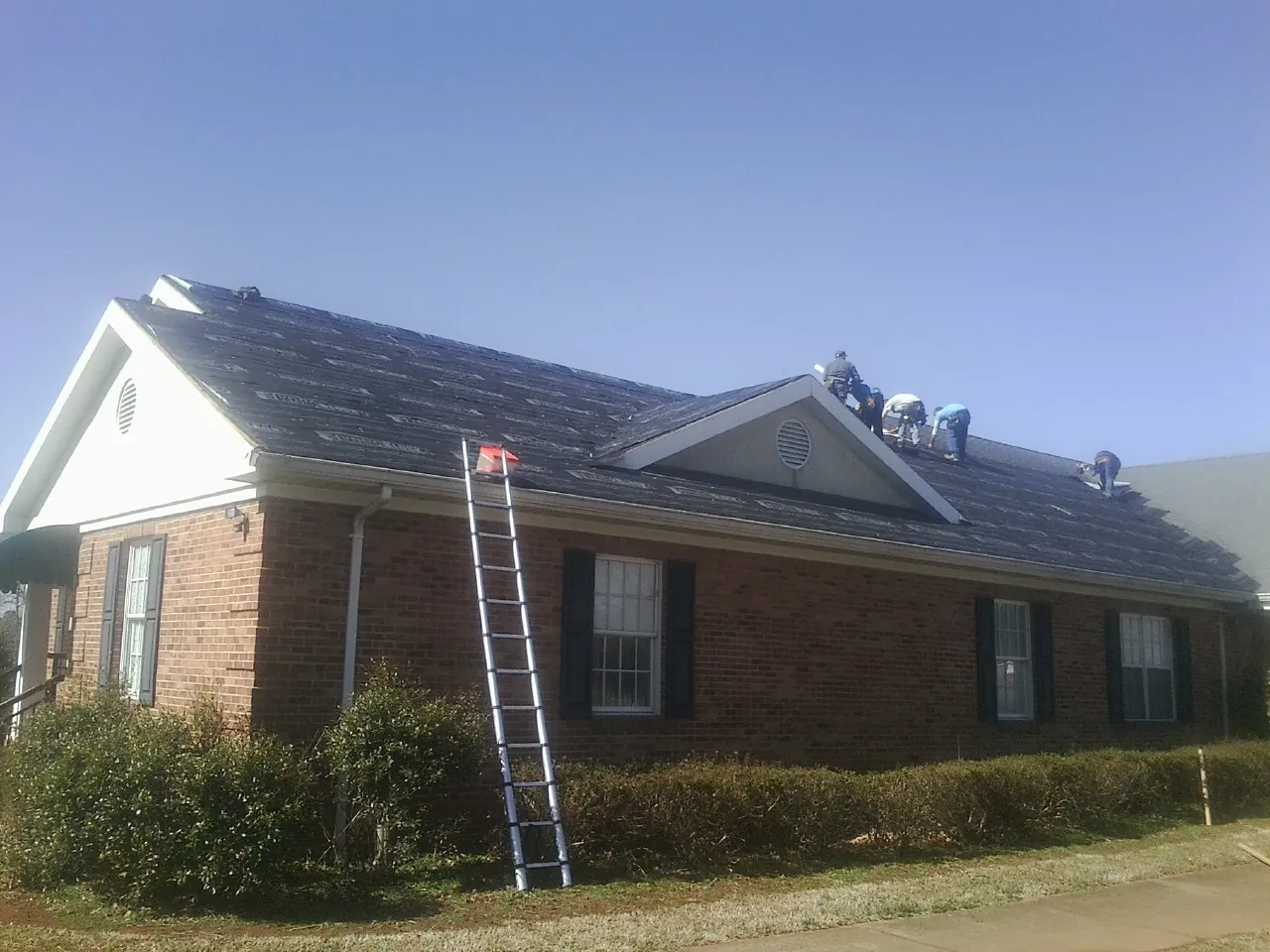 Crew tearing shingles off a brick ranch roof with a ladder set — Emergency Roof Repair work by our crew in Leland